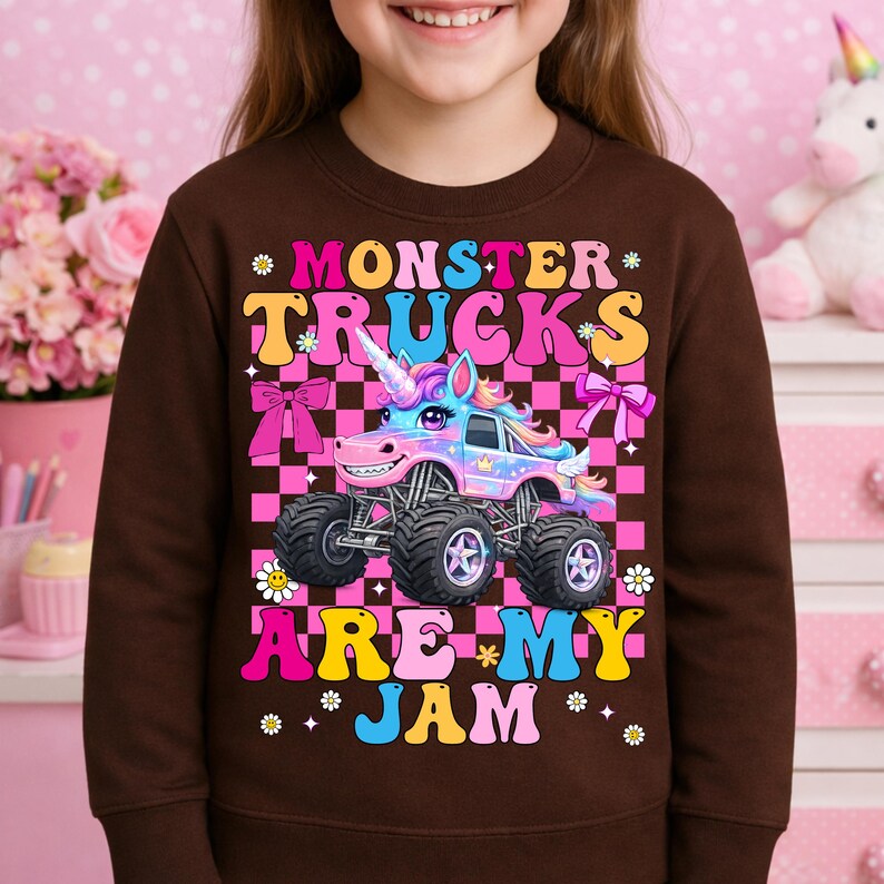 May include: A brown sweatshirt featuring a colorful graphic of a monster truck designed as a unicorn. The text "MONSTER TRUCKS ARE MY JAM" is displayed in a playful font. The background is a pink and black checkered pattern.
