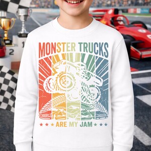 May include: White sweatshirt with a colorful graphic that says "MONSTER TRUCKS ARE MY JAM." The design features a monster truck and several smaller car illustrations in a retro style. The sweatshirt is worn by a person.
