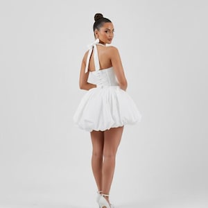 May include: A white halter dress with a bubble skirt. The dress has a fitted bodice and a bow at the neck. The model is wearing white high heels. The background is plain white.