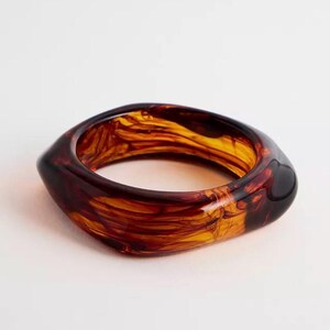 以下が含まれることがあります： 分厚い四角いバングルブレスレット。ブレスレットは、琥珀色、茶色、黄色のマーブル模様の半透明素材で作られています。ブレスレットは滑らかで光沢のある仕上がりで、白い背景に配置されています。