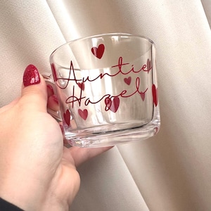 May include: A clear glass mug with a handle, decorated with the words "Auntie Hazel" in red script and red heart motifs. The mug is designed for drinks and is held by a person.