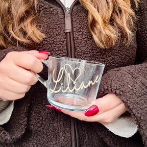 May include: A clear glass mug with a handle, featuring the name "Liliane" in a cursive font with a heart design. The mug is held against a brown fleece jacket.