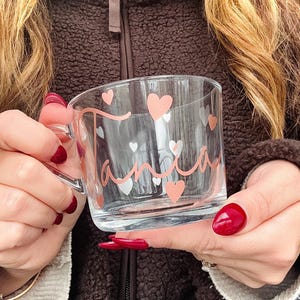 May include: A clear glass mug with a handle, decorated with pink hearts and the name "Tania" in a cursive font. The mug is held by a person with red nail polish.