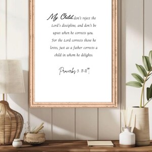 May include: A framed print with a light brown wooden frame. The print features elegant script that reads "My Child... Proverbs 3: 11-12". The text is centered on a white background. The frame is displayed on a wooden shelf.
