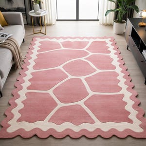 May include: A large, rectangular rug with a pink and off-white design. The rug features a pink central area with an abstract pattern in off-white. The border has a scalloped edge. The rug is placed on a light-colored wooden floor.
