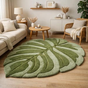 May include: A large, green, leaf-shaped rug with varying shades of green. The rug features a detailed design mimicking the veins of a leaf. It is placed on a wooden floor in a living room setting, with a sofa, chair, and coffee table.