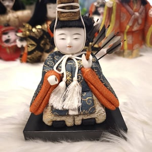 Puede incluir: Una muñeca japonesa con una expresión serena, vestida con un atuendo tradicional con mangas naranjas y un kimono azul y dorado estampado. La muñeca sostiene flechas y está sobre una base negra. Otras muñecas son visibles en el fondo.