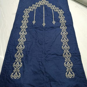 Può includere: Un tappeto da preghiera blu navy con un design ricamato color crema. Il design include una parte superiore ad arco e linee verticali con elementi decorativi. Il tappeto ha una trama trapuntata e una forma a mezzaluna al centro.