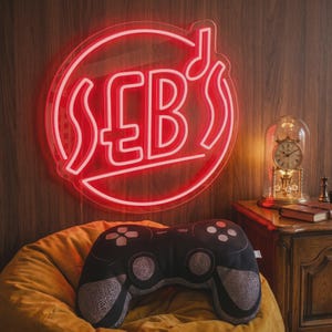 May include: A red neon sign with the text "SEB'S" in a circle, above a black and gray video game controller-shaped pillow. The pillow rests on a yellow beanbag chair. A vintage clock is on a wooden nightstand.
