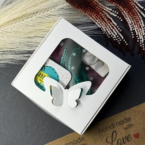 May include: A white square gift box with a butterfly-shaped cutout window, containing turquoise beaded jewelry on a display card. Beside the box are long, dangling earrings featuring a gradient of dark brown to white beads on silver hoops.