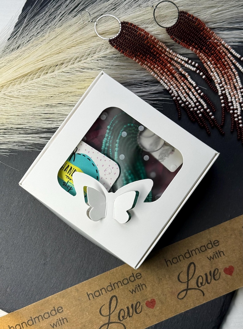 May include: A white square gift box with a butterfly-shaped cutout window, containing turquoise beaded jewelry on a display card. Beside the box are long, dangling earrings featuring a gradient of dark brown to white beads on silver hoops.