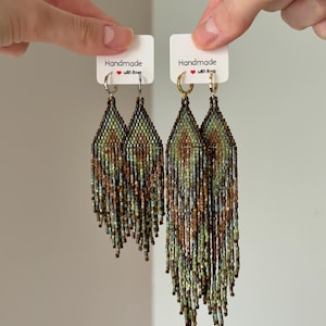 May include: Two pairs of long, diamond-shaped beaded fringe earrings featuring a geometric pattern in shades of green, brown, and metallic bronze. The left pair hangs from silver-tone hoops, while the right pair hangs from gold-tone hoops on white cards.