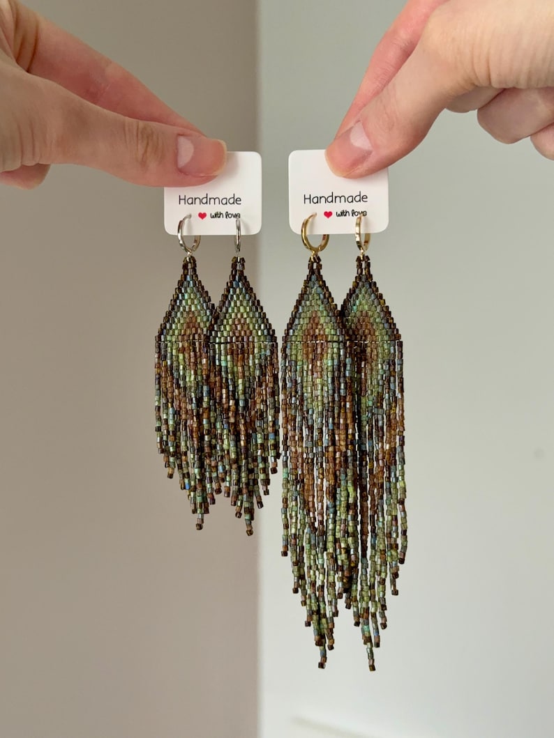 May include: Two pairs of long, diamond-shaped beaded fringe earrings featuring a geometric pattern in shades of green, brown, and metallic bronze. The left pair hangs from silver-tone hoops, while the right pair hangs from gold-tone hoops on white cards.