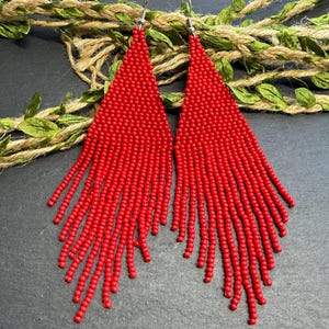 Peut inclure: Une paire de longues boucles d'oreilles triangulaires composées de perles de rocaille rouge vif. Chaque boucle présente un sommet triangulaire perlé solide qui se prolonge par une frange en cascade de brins perlés, avec des crochets en métal argenté.