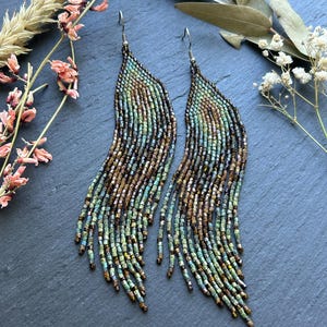 May include: A pair of long, dangling earrings crafted from small, iridescent glass beads in shades of teal, bronze, and copper. The beads are arranged in a curved, feather-like pattern with multiple fringe strands hanging from silver-toned hooks.