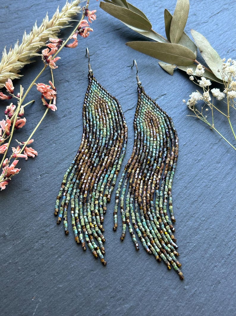 May include: A pair of long, dangling earrings crafted from small, iridescent glass beads in shades of teal, bronze, and copper. The beads are arranged in a curved, feather-like pattern with multiple fringe strands hanging from silver-toned hooks.