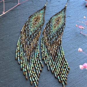 May include: A pair of long, dangling earrings crafted from small, iridescent glass beads in shades of bronze, copper, and teal. The beads are arranged in a curved, fringe-like pattern that tapers toward the bottom, suspended from silver-toned hooks.
