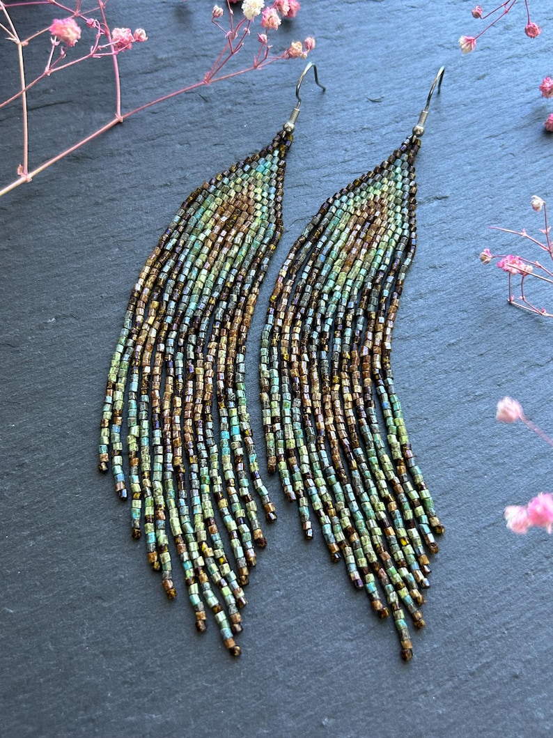 May include: A pair of long, dangling earrings crafted from small, iridescent glass beads in shades of bronze, copper, and teal. The beads are arranged in a curved, fringe-like pattern that tapers toward the bottom, suspended from silver-toned hooks.