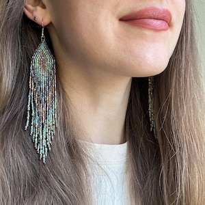 May include: Long, dangling fringe earrings crafted from tiny glass seed beads in a gradient of metallic bronze, muted teal, and soft sage green. The beaded design features a pointed, leaf-like shape at the top that transitions into loose, flowing strands.