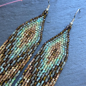 May include: A pair of long, dangling earrings crafted from small, square beads in shades of metallic bronze, copper, and turquoise. The beads are arranged in a geometric pattern, suspended from silver-toned metal hooks that attach at the top.