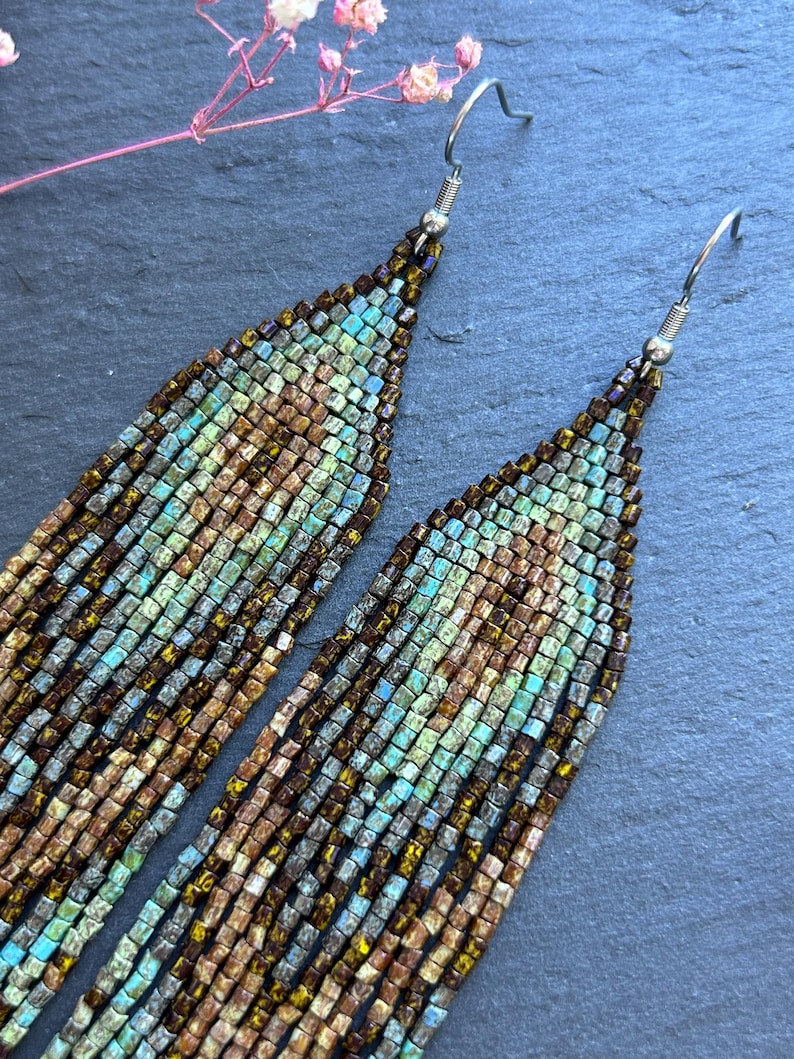 May include: A pair of long, dangling earrings crafted from small, square beads in shades of metallic bronze, copper, and turquoise. The beads are arranged in a geometric pattern, suspended from silver-toned metal hooks that attach at the top.