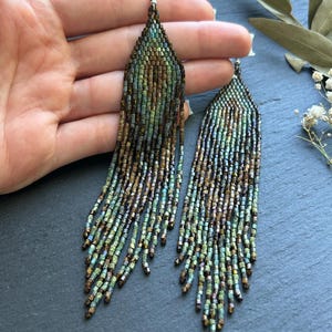 May include: A pair of long, dangling earrings crafted from small, iridescent glass beads in shades of metallic bronze, deep forest green, and teal. The earrings feature a triangular top section that transitions into multiple thin, beaded fringe strands.