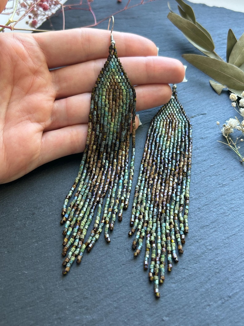 May include: A pair of long, dangling earrings crafted from small, iridescent glass beads in shades of metallic bronze, deep forest green, and teal. The earrings feature a triangular top section that transitions into multiple thin, beaded fringe strands.