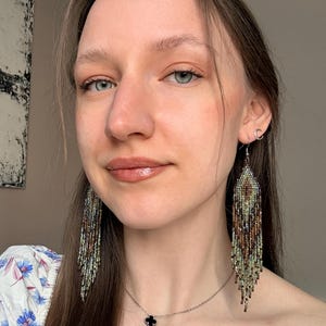 May include: Long, dangling fringe earrings crafted from tiny glass beads in a gradient of earthy tones, including shades of brown, bronze, sage green, and cream. The earrings feature a diamond-shaped top section that tapers into multiple strands.