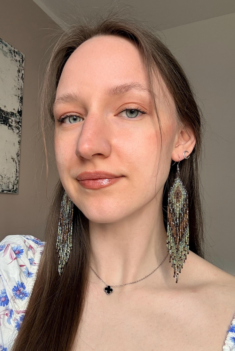 May include: Long, dangling fringe earrings crafted from tiny glass beads in a gradient of earthy tones, including shades of brown, bronze, sage green, and cream. The earrings feature a diamond-shaped top section that tapers into multiple strands.