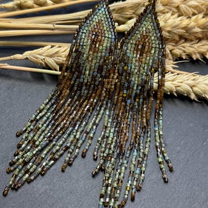 May include: A pair of long, triangular fringe earrings crafted from small glass beads in shades of metallic bronze, muted green, and soft blue. The earrings feature a pointed top section with a geometric pattern and long, dangling beaded strands.