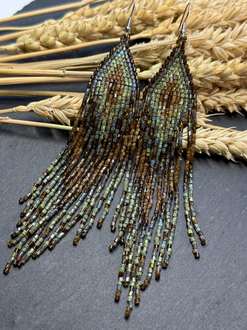 May include: A pair of long, triangular fringe earrings crafted from small glass beads in shades of metallic bronze, muted green, and soft blue. The earrings feature a pointed top section with a geometric pattern and long, dangling beaded strands.