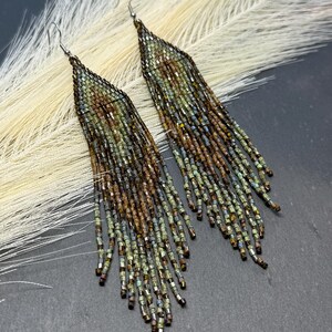 May include: A pair of long, triangular fringe earrings crafted from small glass beads in shades of metallic bronze, olive green, and dark brown. The earrings feature a tapered top section that transitions into multiple dangling beaded strands.
