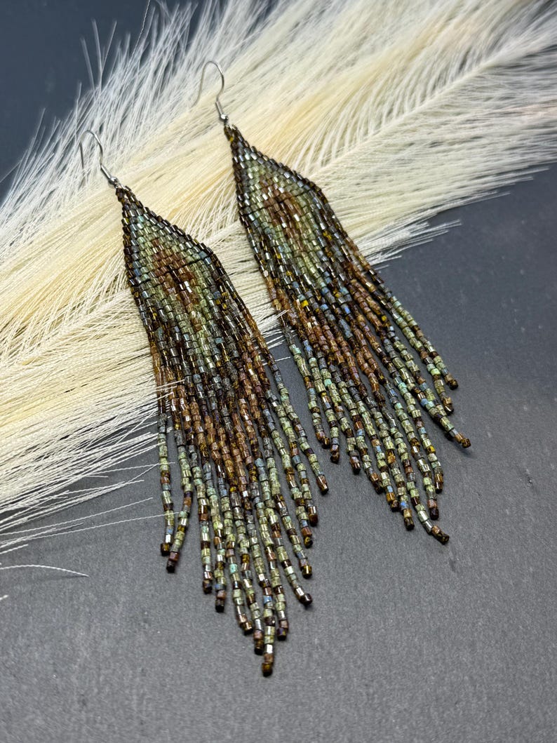 May include: A pair of long, triangular fringe earrings crafted from small glass beads in shades of metallic bronze, olive green, and dark brown. The earrings feature a tapered top section that transitions into multiple dangling beaded strands.