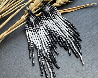 May include: A pair of long, dangling earrings crafted from small black, white, and metallic silver seed beads. The earrings feature a triangular top section with a diamond pattern, transitioning into multiple cascading fringe strands of varying lengths.