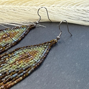 May include: A pair of long, teardrop-shaped dangle earrings crafted from small, multi-colored glass beads in shades of olive green, bronze, brown, and metallic blue. The earrings feature silver-toned metal fishhook ear wires for attachment.
