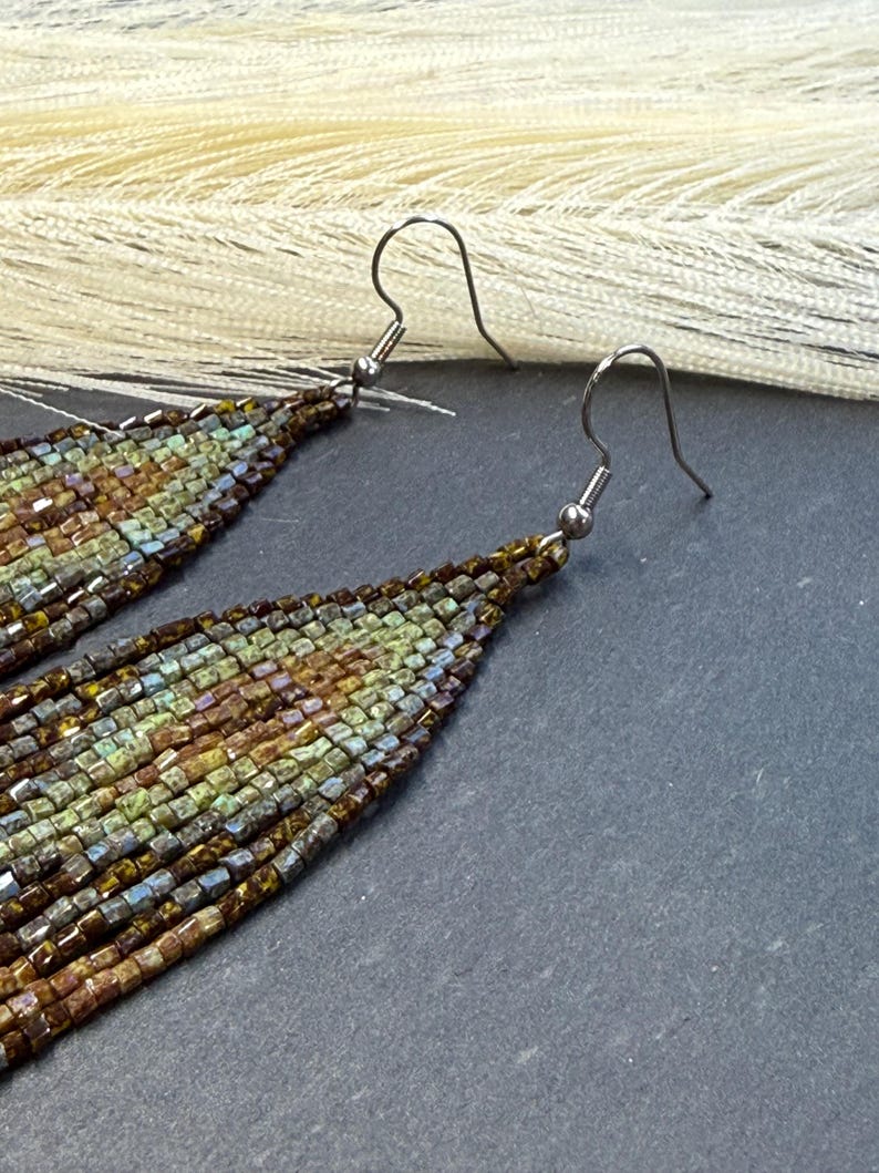 May include: A pair of long, teardrop-shaped dangle earrings crafted from small, multi-colored glass beads in shades of olive green, bronze, brown, and metallic blue. The earrings feature silver-toned metal fishhook ear wires for attachment.