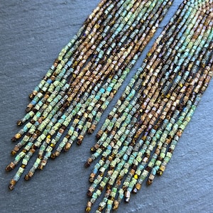 May include: A pair of long, dangling earrings crafted from numerous strands of small, cylindrical beads. The beads feature a gradient color pattern transitioning from metallic bronze and brown tones at the top to shades of teal and light green at the bottom.
