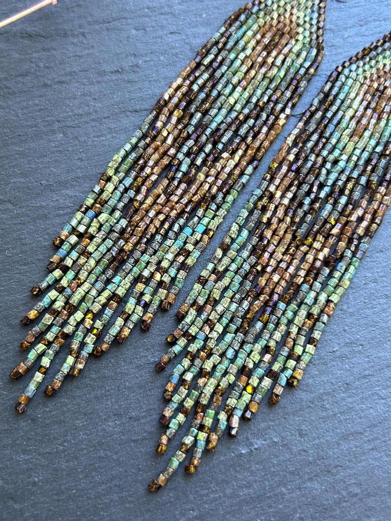 May include: A pair of long, dangling earrings crafted from numerous strands of small, cylindrical beads. The beads feature a gradient color pattern transitioning from metallic bronze and brown tones at the top to shades of teal and light green at the bottom.