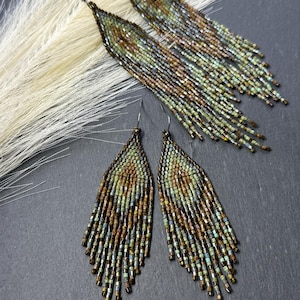 May include: Two pairs of long, triangular fringe earrings crafted from small, iridescent glass beads in shades of bronze, copper, sage green, and muted teal. The beads are arranged in a geometric pattern with dangling fringe strands at the base.
