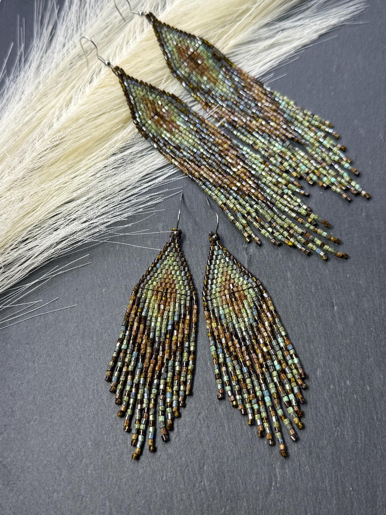 May include: Two pairs of long, triangular fringe earrings crafted from small, iridescent glass beads in shades of bronze, copper, sage green, and muted teal. The beads are arranged in a geometric pattern with dangling fringe strands at the base.