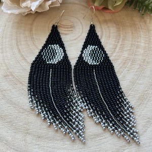 Può includere: Un paio di orecchini a frange nere con perline con un design circolare bianco e argento. Gli orecchini hanno una forma a goccia e pendono da ganci argentati. La frangia è un mix di perline nere e argento.