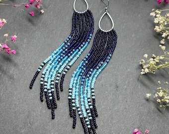 May include: A pair of long, beaded earrings with a teardrop-shaped silver top. The beads transition from dark blue to light blue, creating a gradient effect. The earrings are a stylish accessory.