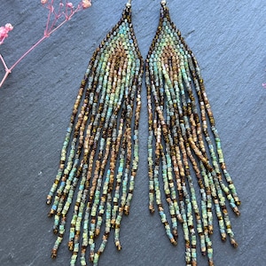 May include: A pair of long, triangular fringe earrings crafted from small glass beads in shades of metallic bronze, teal, and light blue. The earrings feature a geometric pattern that transitions into multiple dangling beaded strands.
