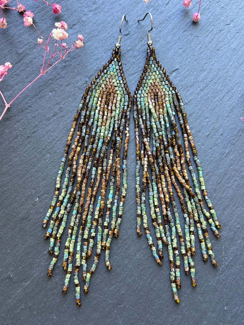 May include: A pair of long, triangular fringe earrings crafted from small glass beads in shades of metallic bronze, teal, and light blue. The earrings feature a geometric pattern that transitions into multiple dangling beaded strands.