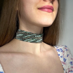 Op de afbeelding: Een kralen choker ketting met een geometrisch patroon. De choker heeft een mix van groene, zwarte, witte en gouden kralen. De ketting wordt om de nek gedragen.