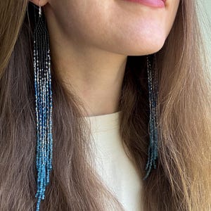 Peut inclure: Longues boucles d'oreilles à franges présentant un motif dégradé passant du noir en haut à l'argent métallique et à diverses nuances de bleu en bas. Les boucles d'oreilles sont composées de petites perles uniformes disposées en brins verticaux.