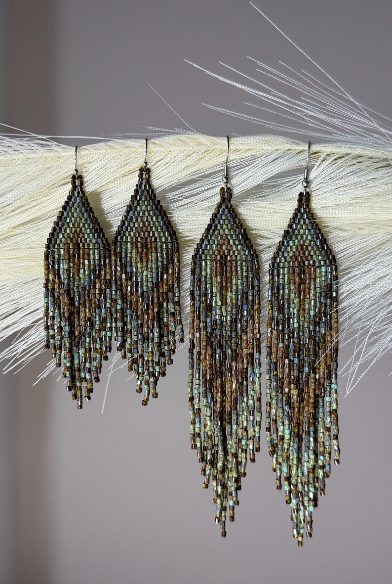 May include: Two pairs of long, teardrop-shaped beaded earrings featuring a woven pattern of metallic brown, bronze, and muted teal glass beads. Each earring has a fringe of dangling beads at the bottom and is attached to a silver-toned metal hook.