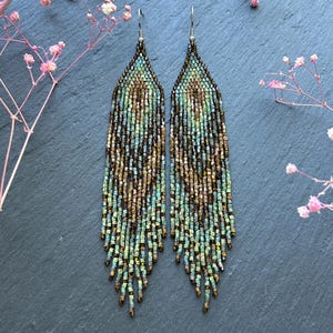 May include: A pair of long, dangling earrings crafted from small, multi-colored glass beads in shades of metallic bronze, forest green, and teal. The beads are arranged in a pointed, triangular pattern at the top, transitioning into a fringe.