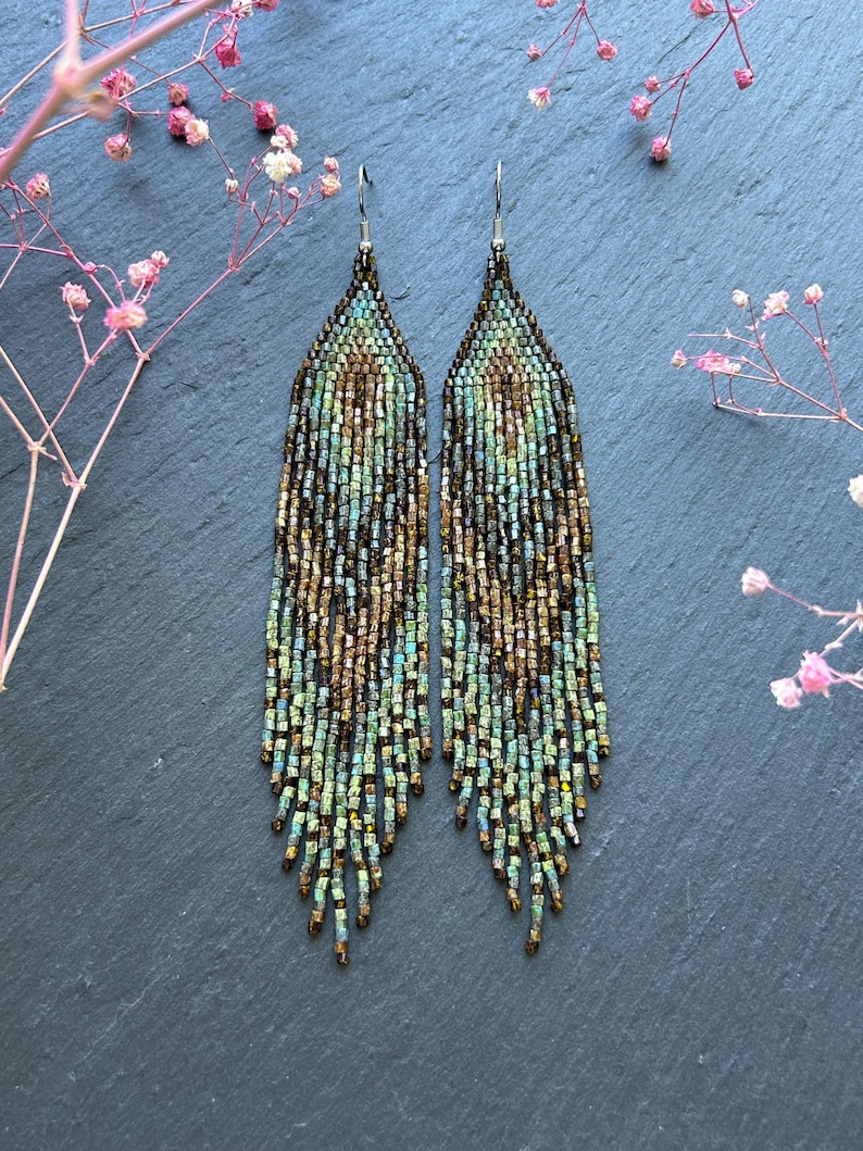 May include: A pair of long, dangling earrings crafted from small, multi-colored glass beads in shades of metallic bronze, forest green, and teal. The beads are arranged in a pointed, triangular pattern at the top, transitioning into a fringe.