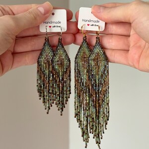 May include: Two pairs of long, beaded fringe earrings featuring a geometric pattern in shades of green, brown, and blue. One pair has silver-toned hoops, and the other has gold-toned hoops. Each pair is attached to a white card labeled Handmade with love.
